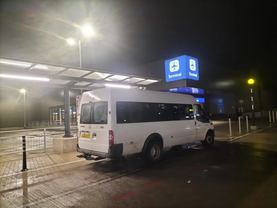 Bristolian Transfers' 16-seat minibus waiting at the Bristol Airport terminal drop-off point at night